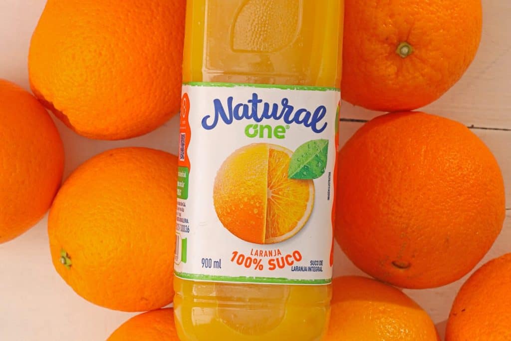 Texto Alternativo - No centro da imagem há uma embalagem do suco Natural One de laranja. No fundo, diversas laranjas.