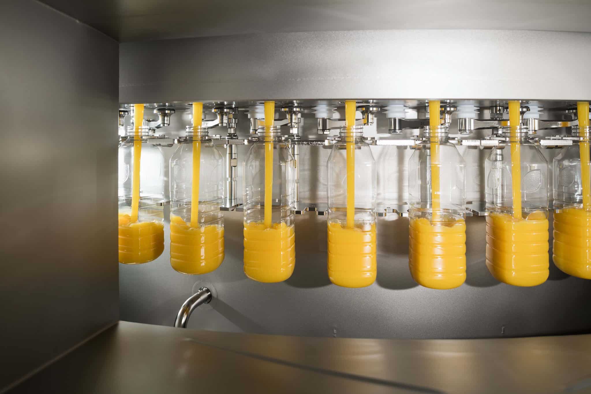 Texto alternativo: Na imagem há cinco garrafas de Natural One sendo abastecidas com sucos de laranja na fábrica.
