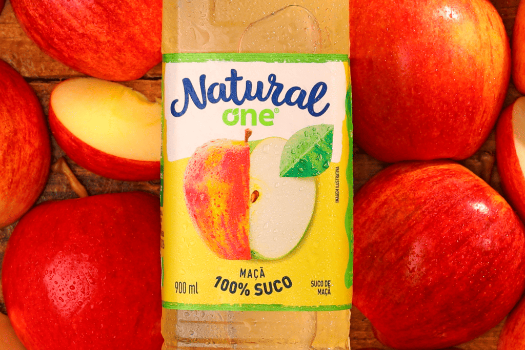 Na imagem há várias maçãs de fundo e ao centro da imagem um suco Natural One de maçã.