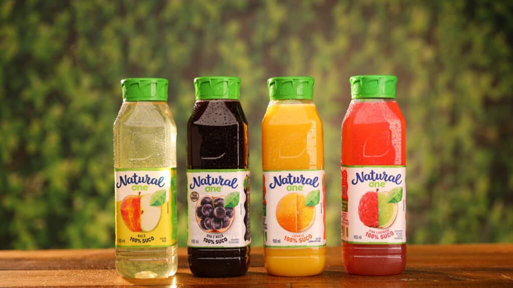 Na imagem há quatro sucos de Natural One de 900ml: maçã, uva, laranja e pink lemonade.