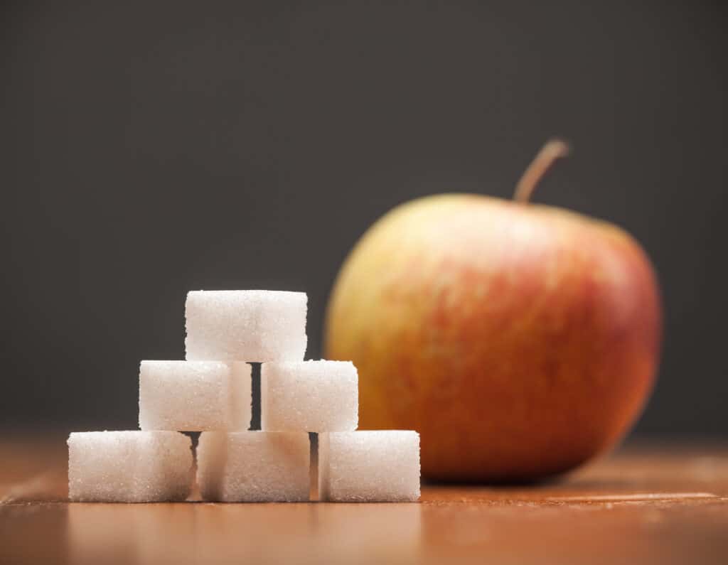 Texto alternativo: Na imagem há seis cubos de açúcar e ao fundo uma maçã vermelha