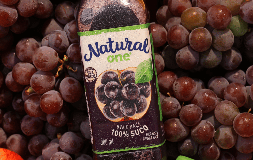 Texto Alternativo: Na imagem há diversas uvas de fundo e como ponto focal o suco de uva da Natural One