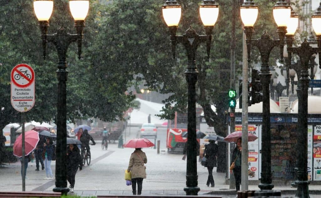 Na imagem há um rua, com iluminação ligada, árvores ao redor, pessoas com guarda-chuva e nevoa