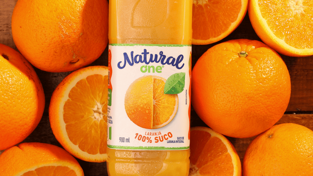 Texto alternativo: Na imagem há uma garrafa de suco de laranja da Natural One. No fundo, há dezena de laranja.