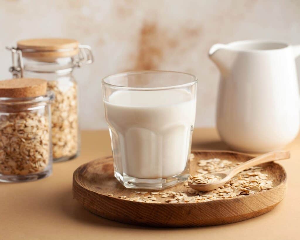 Texto alternativa: Na imagem há um copo de leite (lactose), em recipiente de vidro, no centro da mesa. Ao lado, potes de aveia