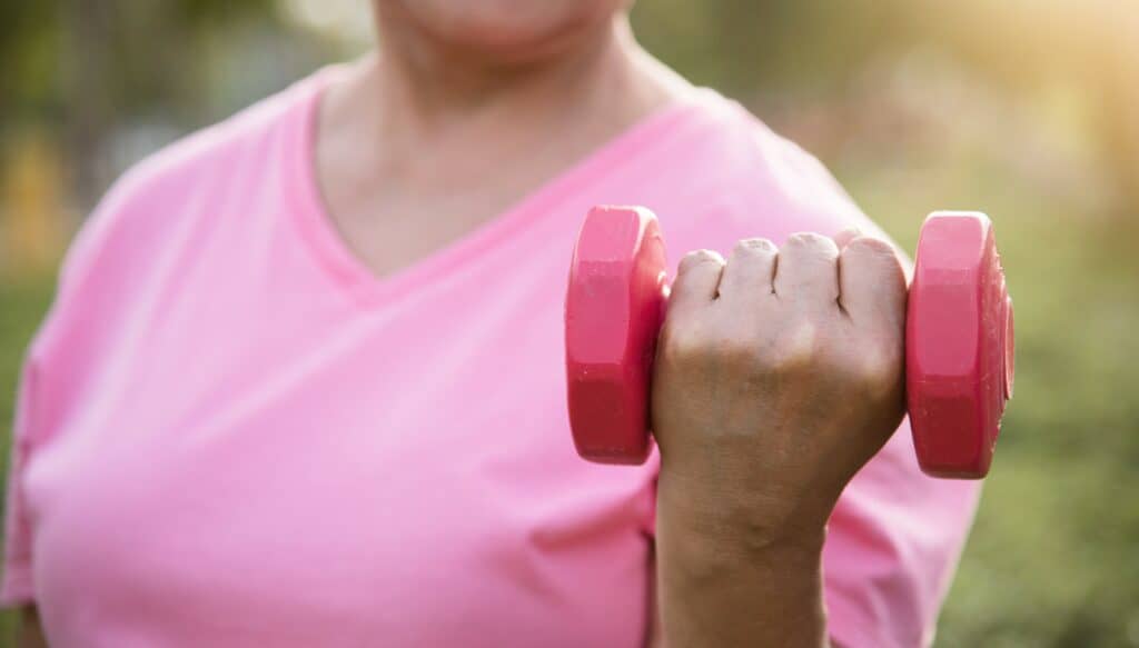 Texto alternativo: mulher segurando peso de academia. Mostra que a atividade física pode te fortalecer no processo de câncer de mama