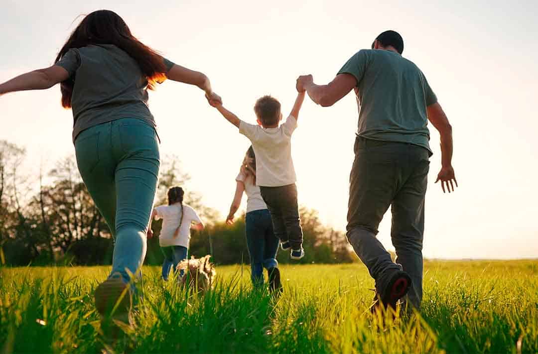 Na imagem há um homem e uma mulher segurando uma criança pelos braços enquanto pulam juntos em uma grama verde, a família também é um ponto chav da saúde mental