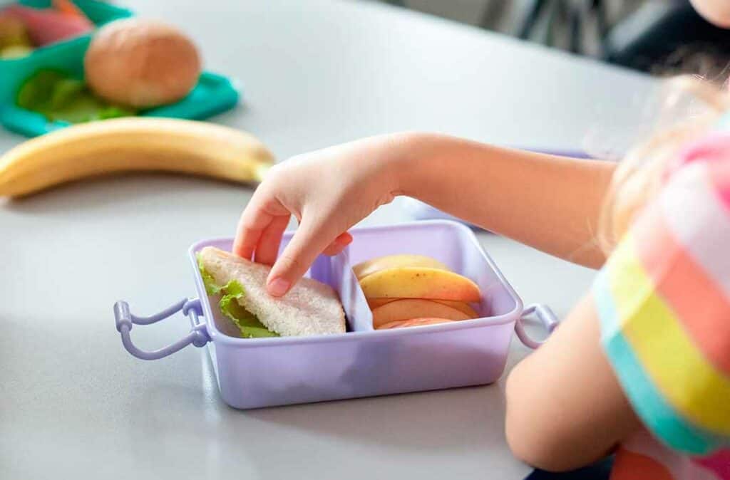 Texto alternativo - Na imagem há uma mão de uma criança colocando frutas em uma lancheira