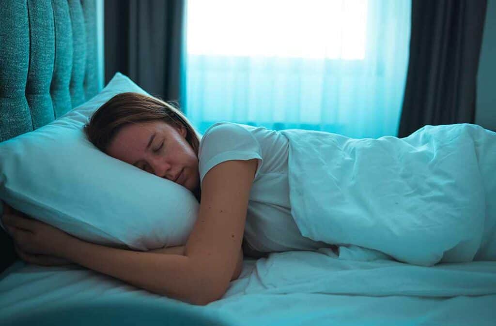 Texto alternativo - na imagem há uma mulher deitada e dormindo em uma cama de lençóis brancos