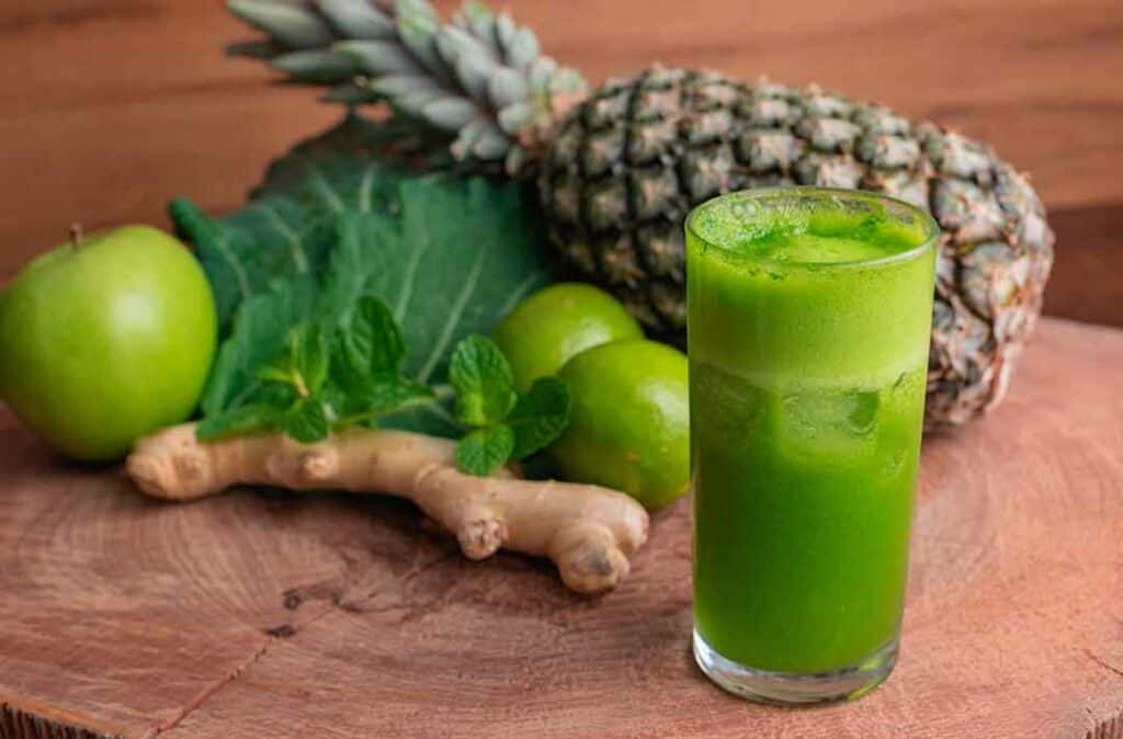 Texto alternativo - Na imagem há um copo de suco verde detox, ao lado um abacaxi, couve e gengibre