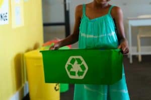 Texto alternativo: na imagem há uma menina de vestido verde, segurando uma caixa indicada como reciclável, indicando a educação ambiental