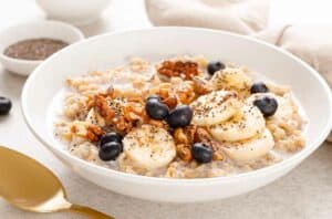 Texto alternativo: oatmeal, com pedaços de banana, mirtilos e lascas de castanha em uma tigela branca com uma colher dourada aparecendo ao lado