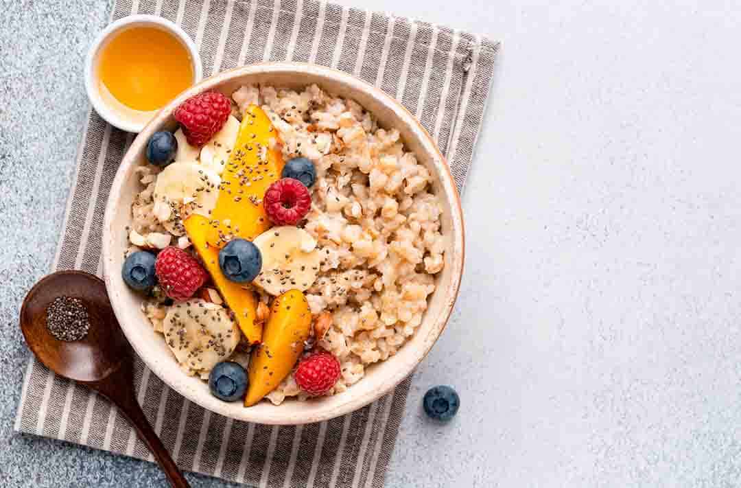 Texto alternativo: oatmeal com diversos toppings, como: banana, mirtilo e framboesa. Também acompanha uma colher ao lado, em cima de uma toalha pequena listrada