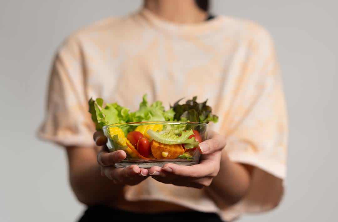 Texto alternativo: uma moça segurando um bowl de vidro que contém folhas de alface e algumas verduras, frutas e legumes