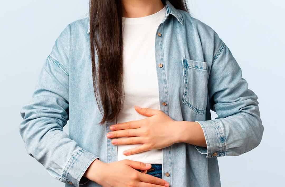 Texto alternativo: uma moça de cabelo moreno e longo, com uma camiseta branca e uma camisa azul clara, com as duas mãos na barriga