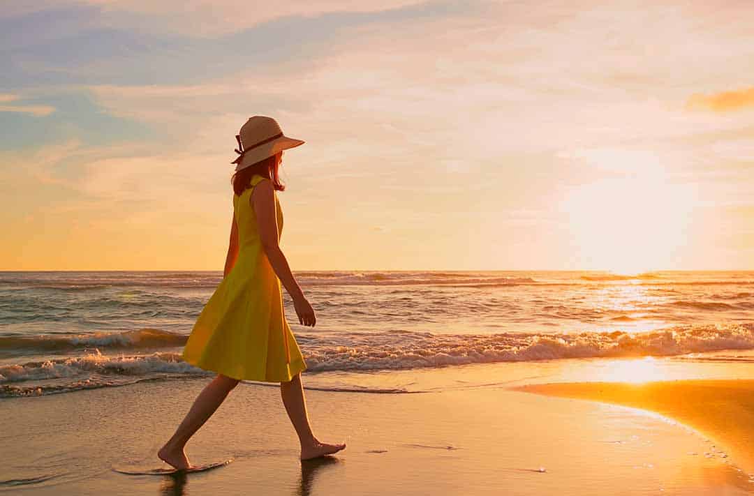 Texto alternativo: uma moça de vestido amarelo, descalça e usando um chapéu, caminhando na areia da praia no pôr do sol. 