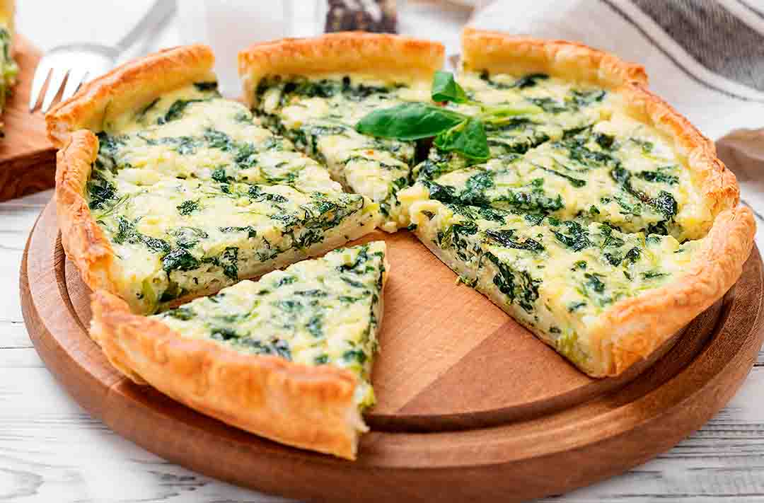 Texto alternativo: uma torta de espinafre com ricota cortada em 8 pedaços, como uma pizza. Está sob um suporte de madeira. Ótima opção para um jantar saudável