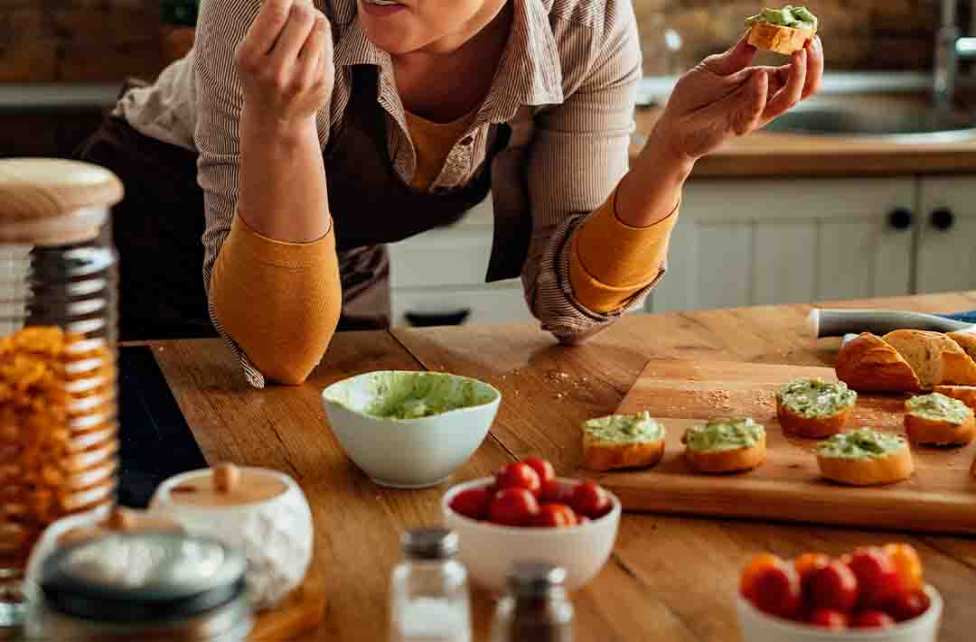 Texto alternativo: uma moça apoiada em uma mesa de madeira que contém diversos tipos de alimentos saudáveis, como frutas, legumes, torradas e patês