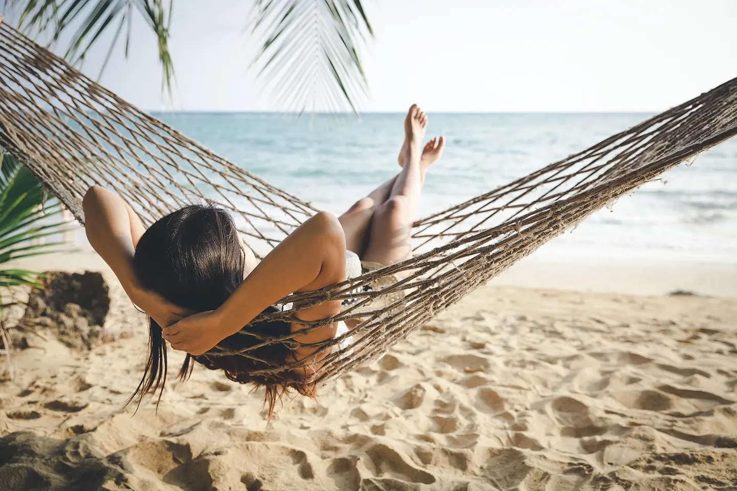 Texto alternativo: uma mulher, deitada em uma rede, na praia, de frente para o mar