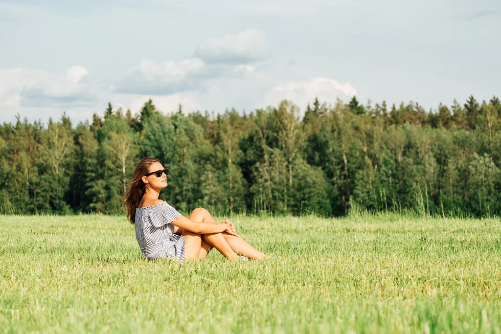 Texto alternativo: na imagem vemos uma mulher sentada na grama, sozinha, e ao fundo uma floresta